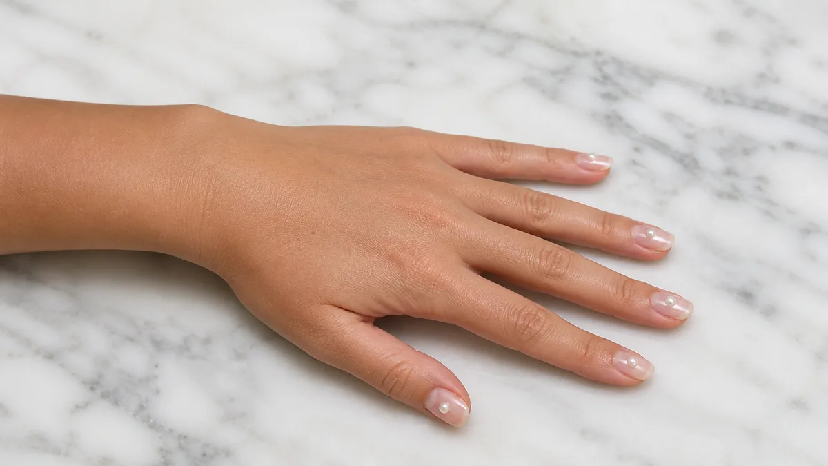 Single left hand with medium oval nails each bearing a single pearl bead on clear gel base palm down on white marble surface
