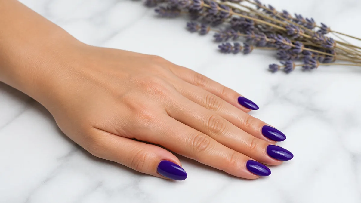 Single left hand with medium almond nails in royal purple crème gloss resting palm down on white marble with dried lavender in background
